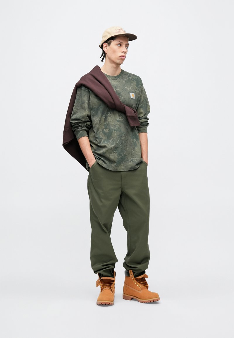 Young man wearing a beige cap, camo long-sleeve shirt, olive pants, brown boots, with a dark sweatshirt draped over his shoulders, standing with hands in pockets.