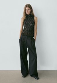 Zwarte kanten mouwloze top met een hoge hals, gecombineerd met zwarte broek met wijd uitlopende pijpen. De outfit heeft een textuurontwerp en een ontspannen pasvorm.