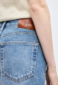 Άτομο που φοράει ανοιχτό μπλε τζιν τζιν με καφέ δερμάτινο ταμπελάκι Calvin Klein στη μέση και μπεζ ριμπ τοπ.