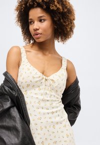 Γυναίκα με σγουρά μαλλιά, ντυμένη με λευκό φόρεμα με λουλούδια και μαύρο δερμάτινο μπουφάν το οποίο κρέμεται από τους ώμους της, κοιτώντας αριστερά σε λευκό φόντο.