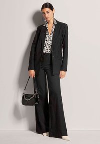 Schwarzer Nadelstreifenanzug mit taillierter Blazerjacke und weit geschnittenen Hosen, kombiniert mit einer gemusterten Bluse. Umfasst eine schwarze glänzende Handtasche und cremefarbene Absätze.