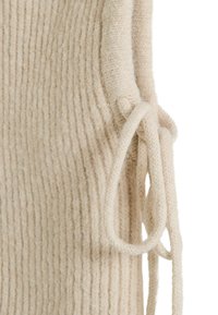 Primer plano de tela beige de punto con un detalle de lazo anudado a un lado sobre un fondo blanco.