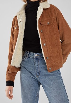 Person iført en brun kordfløjlsjakke med hvid fleeceforing, sort rullekrave og blå højtaljede jeans, der står mod en neutral baggrund.