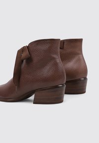 Botas de tobillo de cuero marrón con un acabado texturizado, que presentan un detalle de lazo atado y un diseño de tacón de madera apilada.