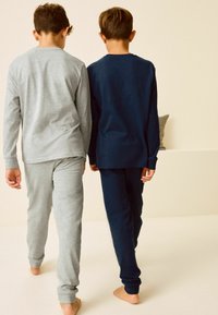 Kaksi lasta kävelee poispäin, päällään harmaat ja tummansiniset pitkähihaiset pyjamat. Kangas on pehmeää, hihansuut ovat tiukat ja malli on rento. Paljaat jalat näkyvät.