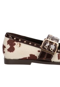 Zapato plano con estampado de vaca, correa de charol marrón, hebilla dorada y acentos con tachuelas. Parte superior de suave textura con patrones contrastantes en marrón y crema.
