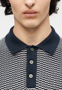 Jeune personne portant un polo en maille bleu marine et blanc avec trois boutons gris et une boucle d'oreille à l'oreille gauche.