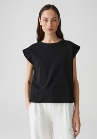 Zwarte katoenen t-shirt met ronde hals en korte kapmouwen met oogjesdetails. Gemaakt in combinatie met witte op maat gemaakte broeken. Soepele textuur.