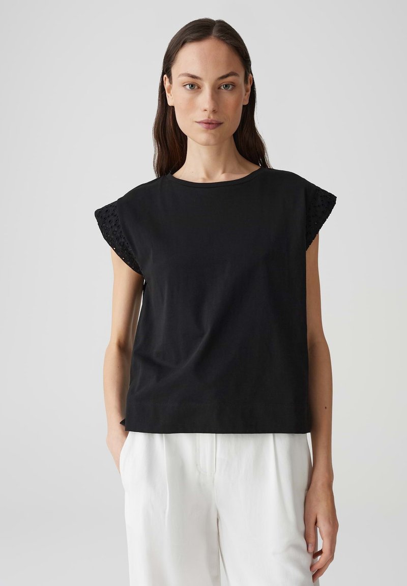 Zwarte katoenen t-shirt met ronde hals en korte kapmouwen met oogjesdetails. Gemaakt in combinatie met witte op maat gemaakte broeken. Soepele textuur.