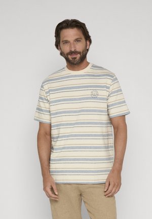 Mand iført beige og blåstribet kortærmet skjorte og lysebrune bukser, stående foran en ensartet lys grå baggrund.