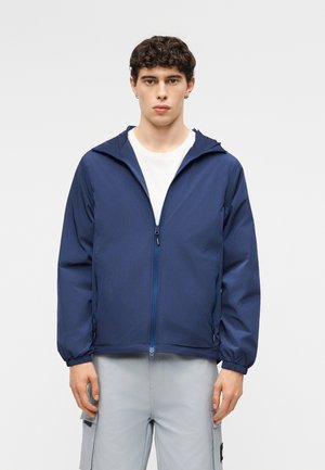 Młody mężczyzna w granatowej kurtce z kapturem na zamek, na białej koszulce i jasnoszarych spodniach typu cargo, stojący na jednolitym tle.