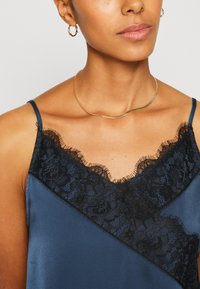 Satin top s dubokim plavim tonom i crnim čipkastim detaljima. Ima V-izrez i uske naramenice. Model nosi zlatne kružne naušnice i jednostavnu ogrlicu.