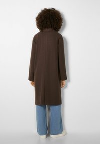 Persona con capelli ricci che indossa un lungo cappotto marrone, jeans a gamba larga azzurri chiari e scarpe bianche, in piedi di fronte a un muro bianco.