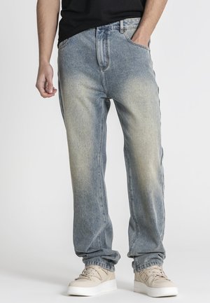 Hombre con jeans vaqueros azules desgastados de corte holgado, zapatillas beige y camiseta negra, de pie frente a un fondo blanco liso.