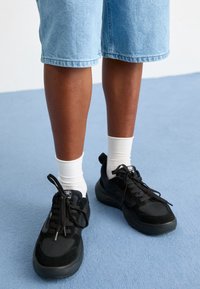 Baskets de sport noires avec un dessus en suede texturé et en mesh, lacets plats, semelle en caoutchouc, portées avec des chaussettes blanches mi-mollet et un short en denim.