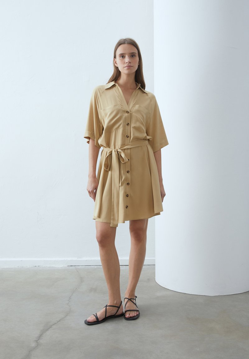 Abito camicia color khaki con maniche corte, apertura frontale con bottoni e cintura coordinata. Tessuto morbido e leggero con due tasche sul petto. Sandali neri.