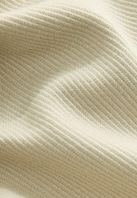 Beige neulekangas, jossa on krooninen rakenne, näyttää pehmeitä laskoksia ja hellävaraisia aaltoilumalleja. Pintakuvio näyttää tasaiselta ja johdonmukaiselta.