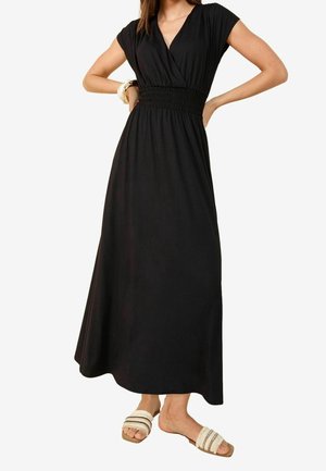 Kobieta ubrana w czarną, bezrękawnikową sukienkę maxi z marszczonym pasem, w połączeniu z białymi płaskimi sandałami o fakturowanej powierzchni oraz złotymi bransoletkami.