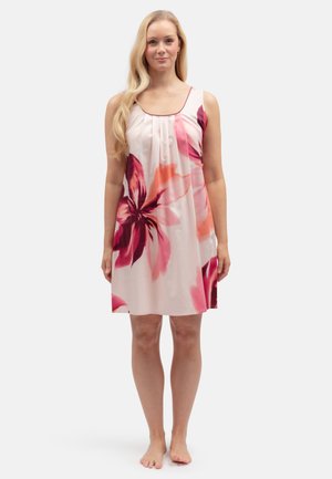 Frau steht barfuß und trägt ein ärmelloses rosa Kleid mit großem roten und orangefarbenen Blumenmuster auf hellem Hintergrund.