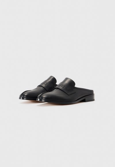Ett par svarta läder loafers utan snörning med låga klackar och märkta remmar över toppen, placerade mot en enkel vit bakgrund.