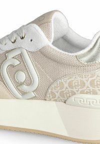 Beige tennari, jossa on teksturoituverkkokangas yläosassa, valkoiset yksityiskohdat, pehmustettu kaulus ja kuvioitu sivupaneeli. Varustettu paksulla valkoisella pohjallisella.