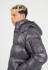 Pufferjack in metallic grijs, met een capuchon, ritszakken en een doorgestikt ontwerp met een glanzende afwerking en zichtbare stiksels.