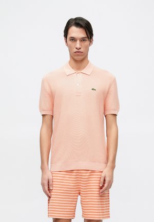 Polo shirt pêche clair en tissu texturé avec des manches courtes, arborant un logo brodé vert. Porté avec un short rayé orange et blanc.