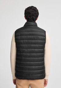 Svart pufferväst med ett kviltat mönster, hög krage och ärmlös design. Tyget verkar slätt och lätt, och ger bra isolering.