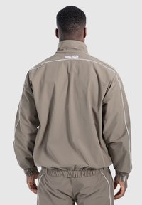 Giacca marrone chiaro con colletto alto, polsini elastici e vita elasticizzata. Presenta profili riflettenti bianchi e il logo "SMILODOX" sul retro.