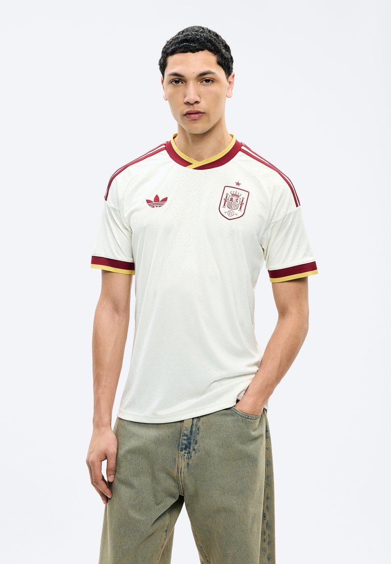 Νεαρός άνδρας με λευκή φανέλα της εθνικής ομάδας ποδοσφαίρου της Ισπανίας, με κόκκινα και κίτρινα περιθώρια, στέκεται με το ένα χέρι στην τσέπη μπροστά σε απλό φόντο.