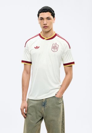 Jeune homme portant un maillot blanc de football d'Espagne avec des bordures rouges et jaunes, debout, une main dans la poche, sur un fond uni.