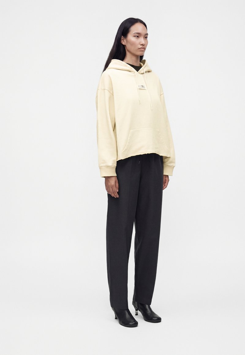 Femme portant un sweat à capuche crème oversized, un pantalon droit noir et des bottines noires à talons, debout devant un fond blanc uni.