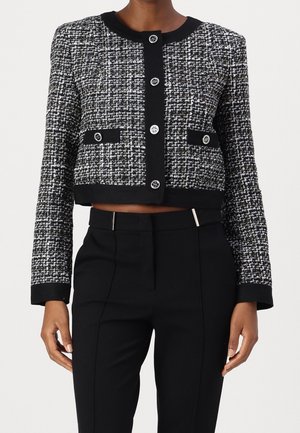 Veste courte en tweed noir et blanc à manches longues et boutons décoratifs, portée avec un pantalon noir taille haute et sur mesure.