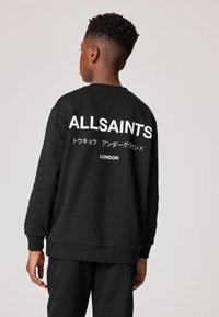 Mladá osoba viděná zezadu, má na sobě černou mikinu s nápisem "ALLSAINTS," japonskými znaky a "LONDON" vytištěnými bílou barvou na zádech.