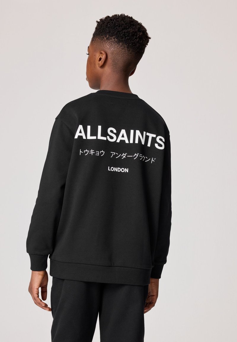 Mladá osoba vidieť zozadu, oblečená v čiernej mikine s nápisom "ALLSAINTS", japonskými znakmi a slovom "LONDON" vytlačenými bielou farbou na zadnej strane.