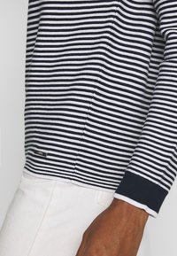 Randig långärmad topp i marinblått och vitt, med ribbade ärmslut, sidofickor och en liten logotyp vid fållen.