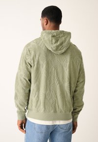 Felpa verde con cappuccio, caratterizzata da motivi in rilievo. Materiale morbido e peluche con polsini aderenti, indossata sopra una camicia bianca e jeans blu.