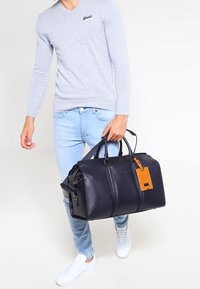 Granatowa torba podróżna z skóry z teksturowanym wykończeniem, podwójnymi górnymi uchwytami, paskiem na ramię i beżową zawieszką bagażową. Posiada podwójne zamknięcie na suwak.