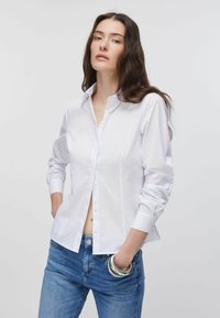 Chemise blanche  à boutons en tissu lisse, avec un col pointu et des manches longues. Associée à un jean bleu et des bracelets argentés.
