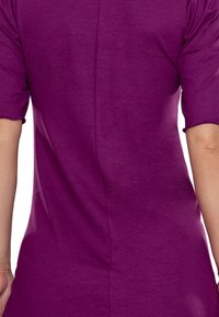 Zbliżenie na osobę w dopasowanej fioletowej bluzce, ukazujące szew tylny i krótkie rękawy. Materiał wydaje się miękki i gładki.