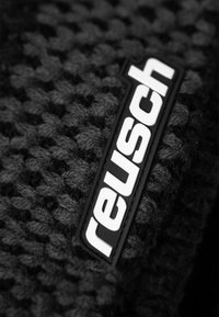 Black textured fabric with a woven pattern, featuring a rectangular Reusch logo in white on a black background.