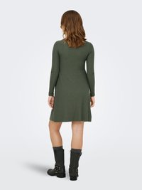 Nainen, jolla on olkapäille ulottuvat ruskeat hiukset, pukeutunut ribbineulottuun, pitkähihaiseen, polvipituiseen vihreään mekkoon ja mustiin keskivartalovarsisaappaisiin harmaiden sukkien kanssa.