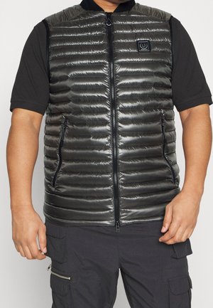 Mies, joka on pukeutunut kiiltävään mustaan tikattuun vetoketjulliseen liiviin mustan lyhythihaisen paidan ja mustien vetoketjutaskullisten cargo-housujen päällä.