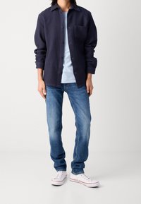 Camisa de botones azul marino oscuro con un patrón texturizado, usada sobre una camiseta azul claro, combinada con vaqueros azules y zapatillas blancas.