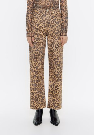 Frau trägt hochgeschnittene, gerade geschnittene Hose mit Leopardenmuster, kombiniert mit passendem langärmligem Leopardenmuster-Oberteil und schwarzen, strukturierten Stiefeletten.