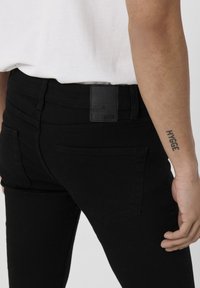 Vaqueros elásticos negros con cintura alta, cinco bolsillos y una etiqueta de cuero rectangular en la parte trasera. Textura suave con un ajuste estilizado.