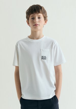Junge mit kurzem braunem Haar trägt ein weißes T-Shirt mit einer vorderen Tasche mit der Aufschrift Scalpers, steht mit den Händen in den Taschen einer dunklen Hose vor einem einfarbigen Hintergrund.
