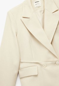 Beżowa, dopasowana marynarka z szerokimi klapami, podwójnym zapięciem na guziki i przednimi kieszeniami. Wykonana z miękkiego materiału o gładkiej fakturze.