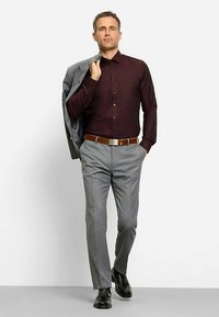 Bordeauxrotes Hemd mit Knöpfen, grauer Blazer über der Schulter, kombiniert mit grauer Hose und schwarzen Schuhen, dazu ein brauner Gürtel.