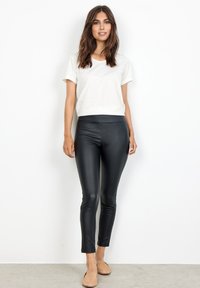 Camicia bianca a maniche corte con scollo a barca abbinata a leggings neri attillati. Il modello indossa delle scarpe basse color sabbia, in piedi contro uno sfondo neutro.